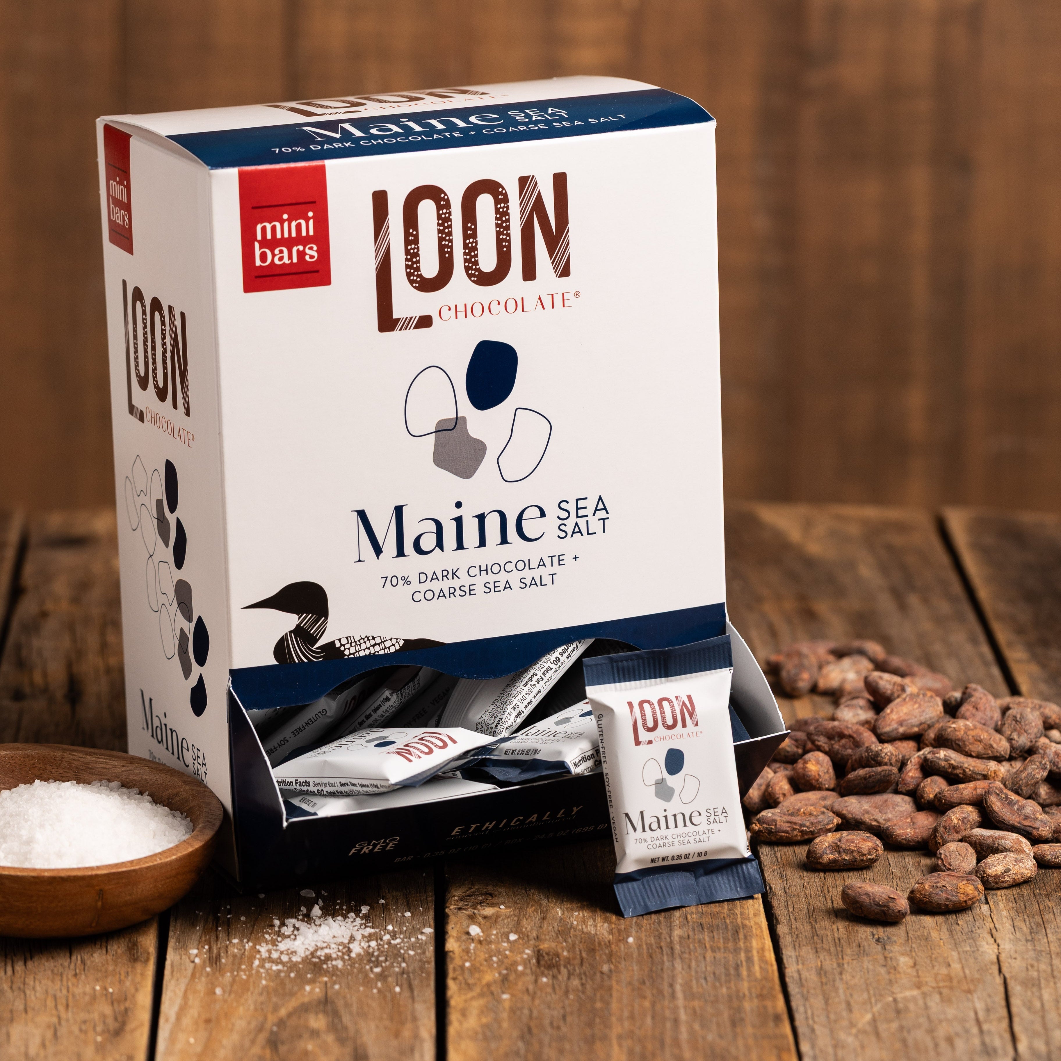 Box of Maine Sea Salt Dark Chocolate mini bars by Loon Chocolate on wooden surface with cacao beans
